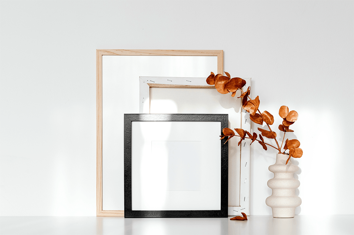 Foto einer weißen Wand die aus dem Lehmfeinputz X-WHITE erzeugt wurde, vor der eine Blumenvase mit  orangenen Blumen, sowie drei verschiedene Bilderrahmen stehen