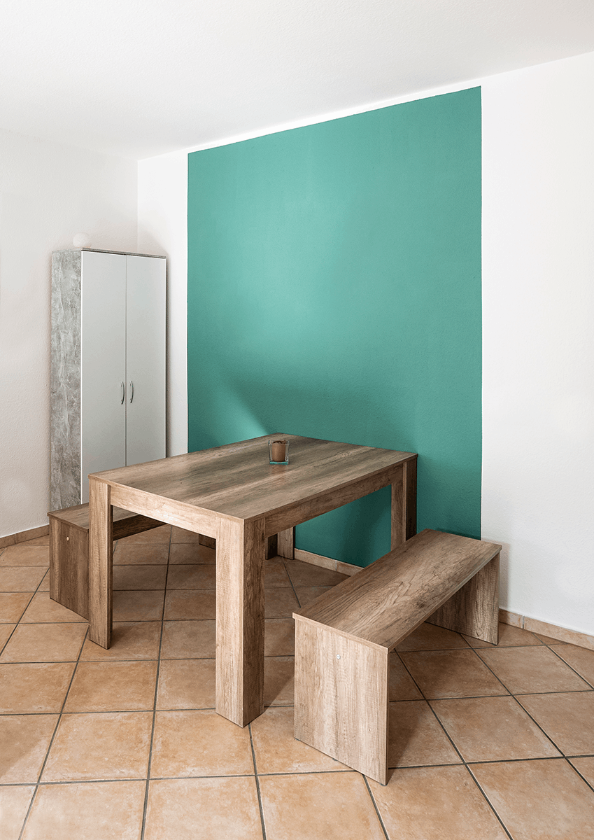 Wand, gestrichen mit der Lehmfarbe I-PAINT und einem grünen Rechteck vor dem auf Fliesen ein Holztisch mit Holbänken steht