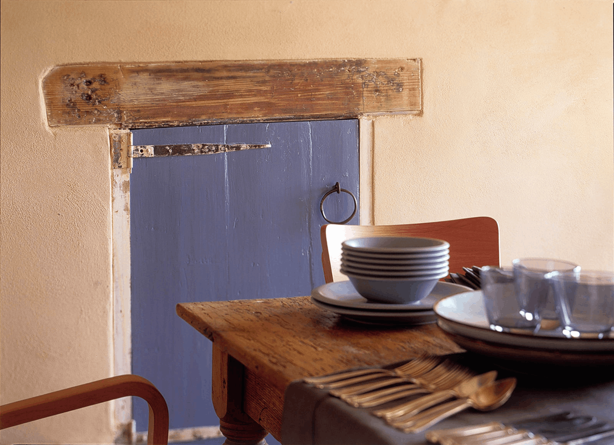Nahaufnahme einer Wand die mit dem Lehmedelputz FINISH erstellt wurde, in der eine lila Holzklappe eingearbeitet wurde und vor der ein großer Holztisch mit Geschirr steht