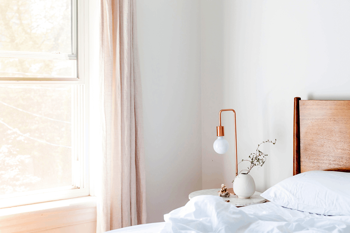 Nahaufnahme eines Schlafzimmerbereiches mit rosefarbenen Vorhängen, sowie einer Nachtischlampe aus Kupfer und einem Holzbett mit weißer Bettwäsche, dessen Wände mit dem Lehmfeinputz X-WHITE verputzt wurden